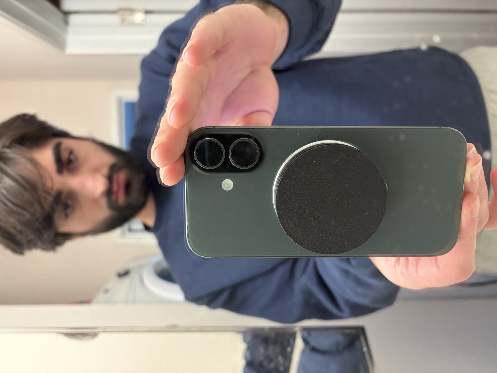 A hand holds up an iPhone from the back. Stuck to the lower middle of the phone is the Linda device: a flat black disc about the size of a silver dollar and as thick as two stacked coins.